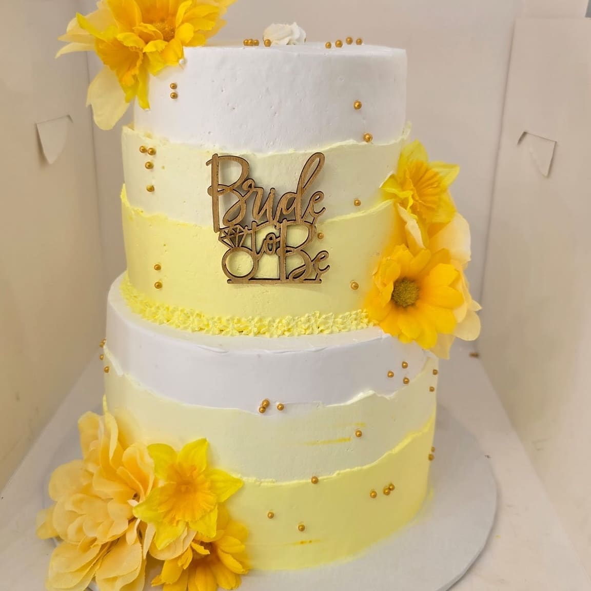 Two tier yellow and white Bride to Be cake with large yellow blooms and a gold script topper