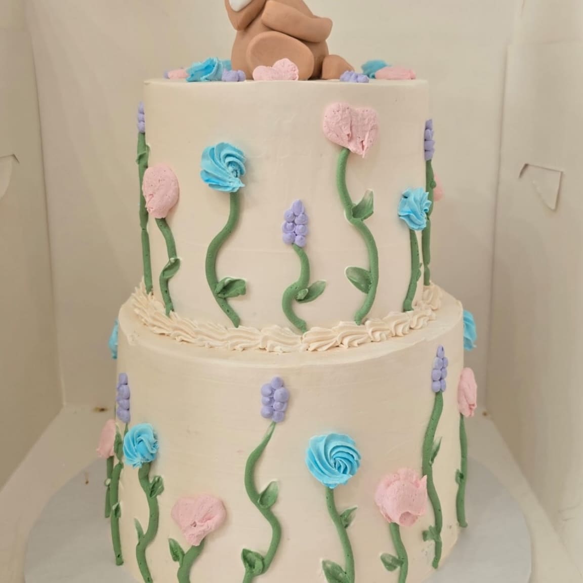 Two tier ivory cake with piped wildflowers in blush, blue and lilac, topped with a sugar fawn