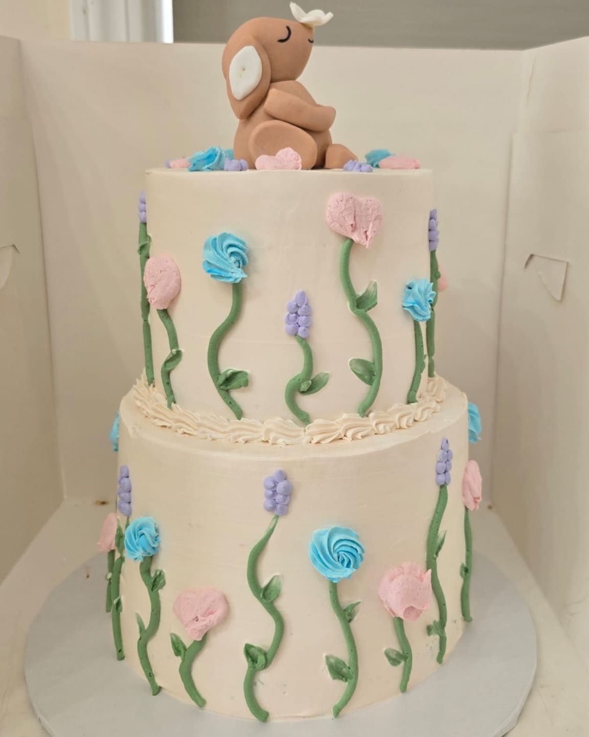 Two tier ivory cake with piped wildflowers in blush, blue and lilac, topped with a sugar fawn