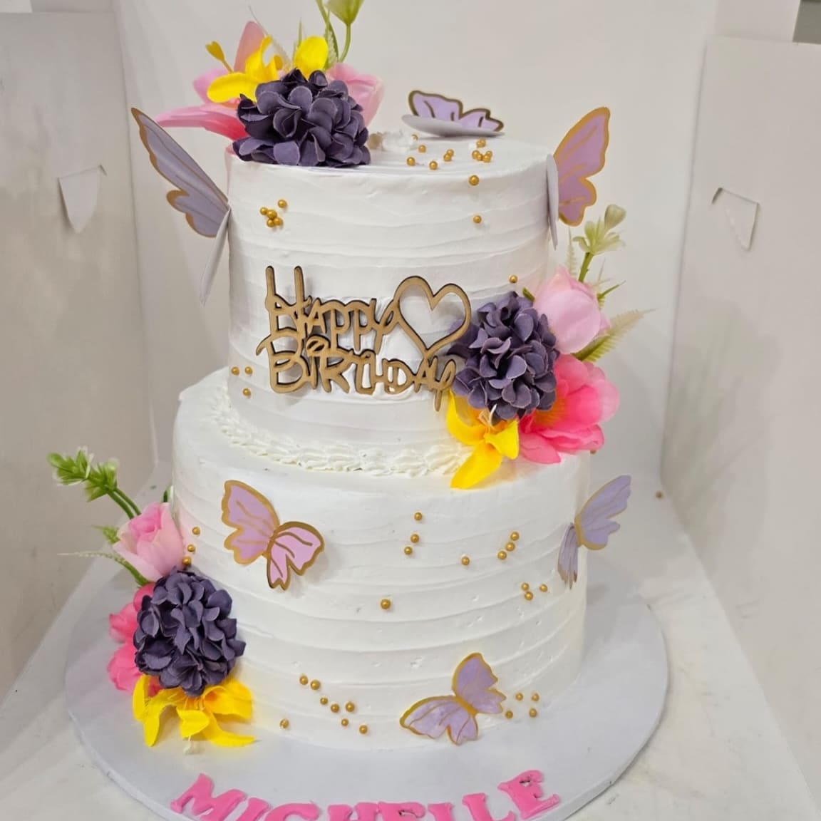 Two tier white birthday cake with purple roses, lilac butterflies, gold pearls and a name piped in pink