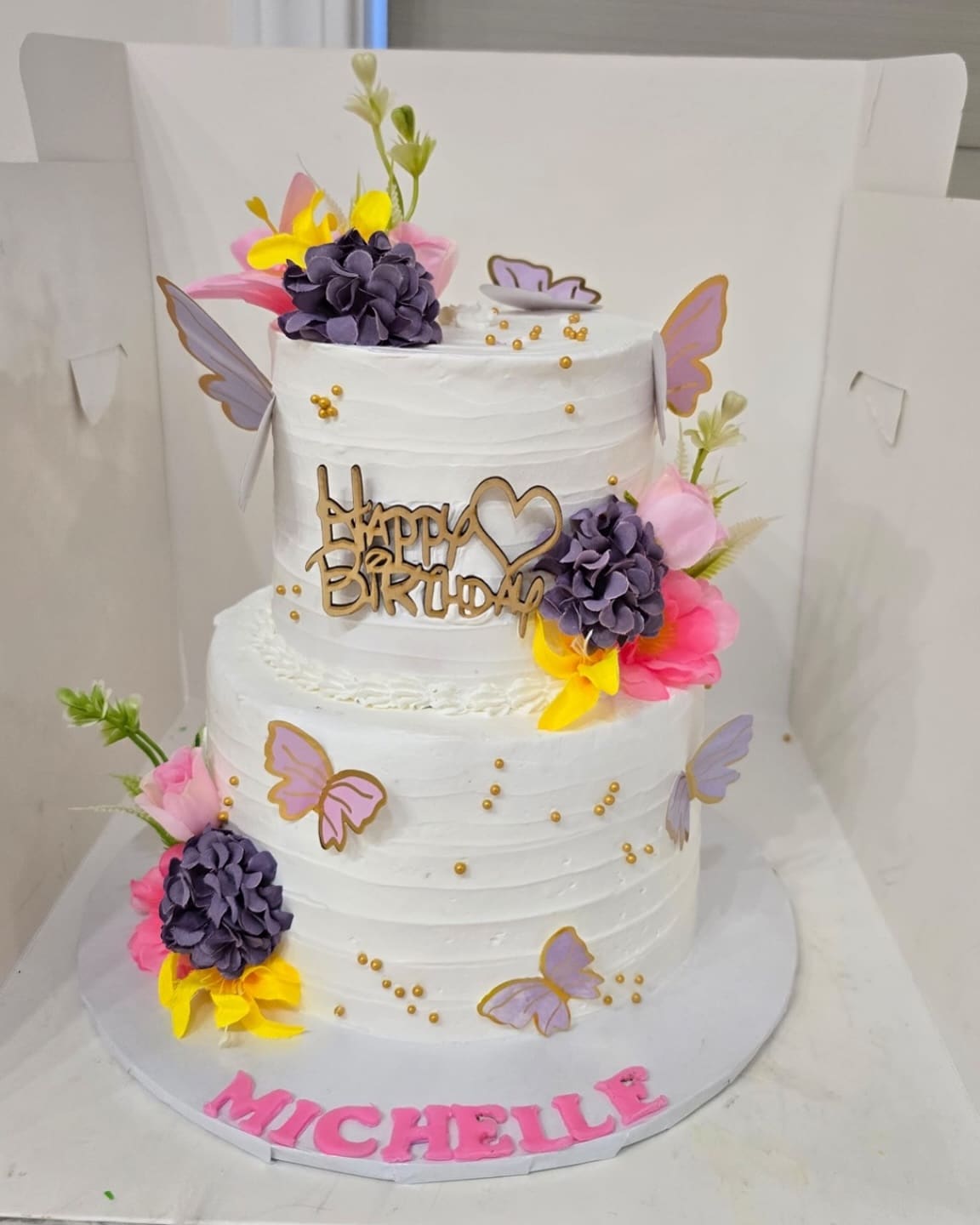 Two tier white birthday cake with purple roses, lilac butterflies, gold pearls and a name piped in pink